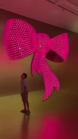 This media file features a young boy standing in front of a large, pink, illuminated bow sculpture. The sculpture is a prominent object, hanging from the ceiling and casting a vibrant pink glow on the surroundings. The boy, dressed in a light-colored t-shirt and shorts, looks up at the bow with a mixture of awe and excitement. His expression and body language suggest he is captivated by the artwork.

The setting appears to be an indoor art exhibition or gallery, indicated by the clean walls and the overhead lighting. The walls are a muted gold color, creating a warm contrast with the bright pink of the sculpture. The floor also reflects the pink light. The overall atmosphere is one of wonder and youthful discovery within an artistic space. The sculpture itself is a significant focal point, detailed with numerous small, bright lights that give it a sparkling, almost dazzling appearance. The time of day is not discernible, but the indoor setting suggests it could be any time. The weather is irrelevant as the scene is indoors.

There is no visible text in this media file. The activity is a child observing and interacting with a large-scale art installation. The boy's posture and gaze convey a sense of wonder and fascination.