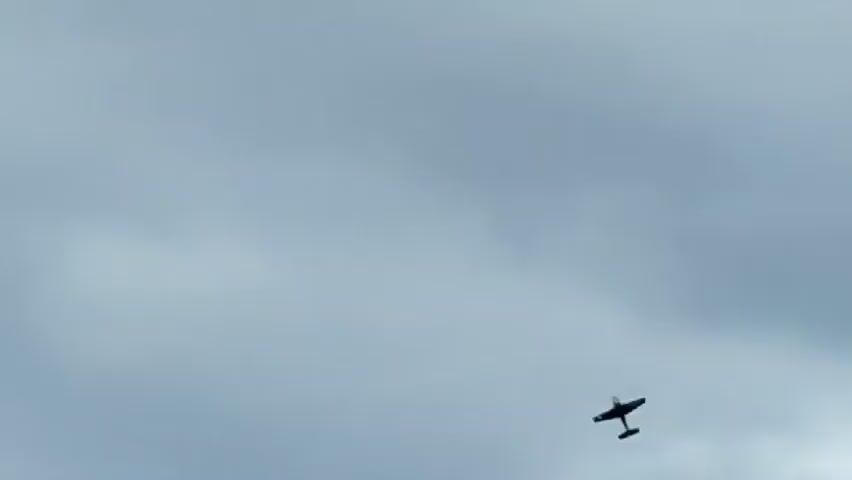 The video shows a dark silhouette of a small airplane against a cloudy, overcast sky. The plane appears to be in flight, and there is a faint white trail behind it, possibly exhaust or condensation. The camera angle is looking upwards, and the plane is a small object in the frame, indicating it is at a considerable distance. The sky is a muted gray, with some lighter patches where the clouds are thinner. The video is short, with the plane remaining visible and in motion throughout the clip.