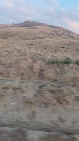 The video is a blurry, horizontal panning shot of a dry, hilly landscape under a pale blue sky with scattered clouds. The foreground is dominated by brown, dry grass and dirt. In the mid-ground, rolling hills covered in similar dry vegetation recede into the distance. A larger, more prominent hill with a rounded peak is visible in the background, appearing tan or light brown. The overall impression is one of a barren or arid environment.