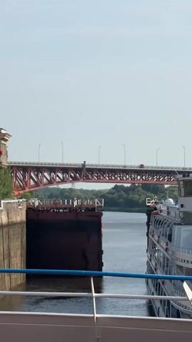 A video shows a river lock with a large ship entering it. The lock gate is open, and the ship is moving slowly into the lock. A large red bridge with an arch spans the river in the background. The sky is clear and blue.