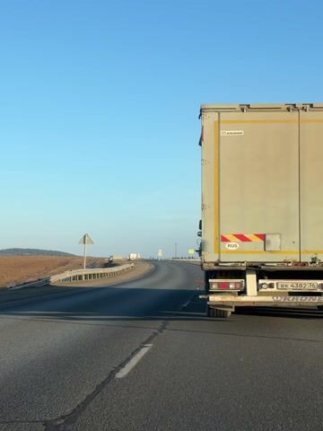 A camera is mounted in a vehicle driving on an asphalt highway on a clear day. The sky is a bright blue. The road is straight and curves gently to the left, with a guardrail and barren brown landscape on the left side. The ground on the right side of the road is covered in snow. The camera initially follows closely behind a large truck with a Krone trailer, identifiable by the logos and "RUS" sticker. The truck slowly pulls ahead. Yellow speed limit signs are visible along the road ahead.