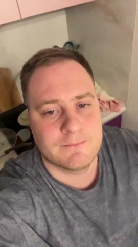 A close-up shot shows a man in his early to mid-30s, wearing a grey t-shirt. He has short brown hair and a light beard, with a tattoo visible on his left inner forearm. He raises his finger and then brings his hand to his face. The background appears to be a kitchen, with pink cabinets and miscellaneous items on the counter. Music plays audibly in the background.