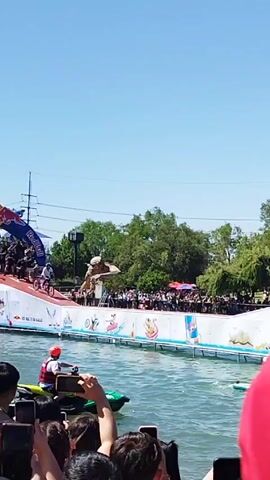 The video shows a man on a bicycle doing a jump off a ramp into the water. The ramp is white and blue and has a Red Bull logo on it. The man is wearing a helmet and is riding a mountain bike. There are people watching from the side, standing behind a white fence. Two people on paddle boards are in the water to the right of the ramp. The water is calm, and the sky is blue. There are trees visible in the background. The video is shot from the perspective of someone in the crowd, with a red hat partially visible in the foreground.