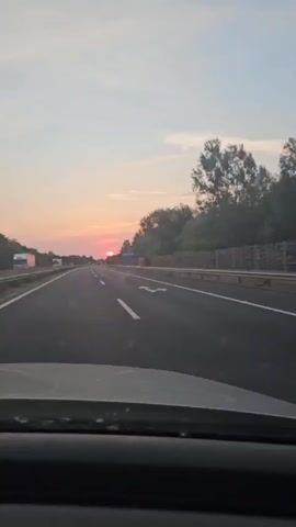 The video shows a first-person perspective of driving on a highway during sunset. The sun is visible on the horizon, casting a warm glow over the sky. Trees and vegetation line the sides of the road, and a highway sign is visible in the distance. The road has multiple lanes and appears to be relatively empty, with only a few other vehicles visible. The dashboard of the car is visible in the foreground.