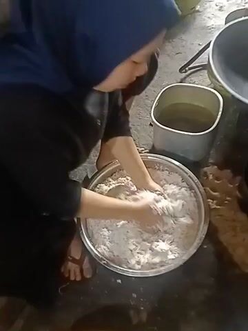 A person wearing a blue hijab and a black abaya is kneeling on the floor and squeezing grated coconut in a large metal bowl. The person is using both hands to knead the coconut, and their feet are also visible near the bowl. A gray rectangular container with water is to the right of the bowl, and other unidentifiable objects are in the background. The floor appears to be dirt or concrete. The action depicted is the process of extracting liquid or milk from grated coconut by squeezing it.