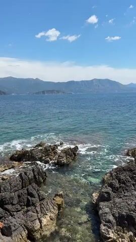 The video shows a rocky coastline. The rocks are dark and jagged, and the water is clear and turquoise. There are small waves crashing against the rocks. In the distance, there are mountains. The sky is blue with some white clouds.