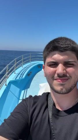 A man takes a selfie outdoors on a boat. He is smiling and looking at the camera. The water and a clear blue sky are visible in the background.