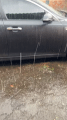 Heavy rain soaks a parked sedan outside a brick building.