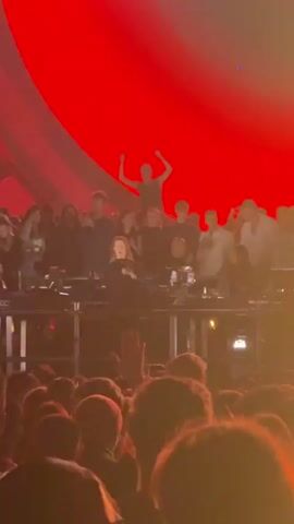 The video shows a crowded indoor music event with a large stage setup. In the foreground, the backs of many people's heads are visible, indicating a large audience. Above them, on the stage, is a raised platform with several individuals. Two people are prominent on this platform: a woman in the center, seated and looking forward, and a man standing behind her with his arms raised in a celebratory gesture. Other individuals are also visible on the platform, some appearing to be part of a DJ setup. The background features a large, curved screen or stage design with red and yellow lighting effects, suggesting a dynamic and energetic atmosphere. The overall impression is a live performance with an engaged audience.