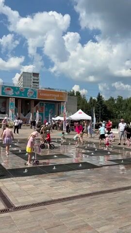 The video shows a sunny day in an outdoor public space with a spray fountain. Several children are playing in the water, some on scooters, and one on a small bicycle. Adults are standing around, some watching the children, others appearing to be browsing stalls set up in the background. Behind the stalls is a building with "КОНЦЕРТНЫЙ ЗАЛ" (Concert Hall) written on it, and trees line the back of the scene. The sky is blue with scattered white clouds.