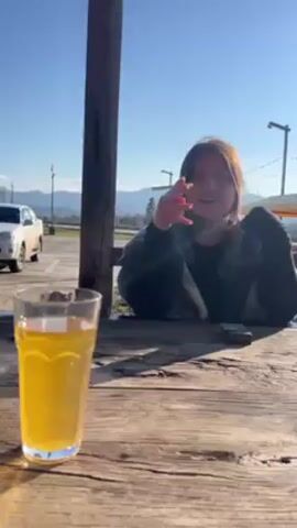 A young woman sits at a weathered wooden picnic table outside, smoking a cigarette and resting her chin on her crossed hands. A full glass of a yellow beverage is in the foreground. She is wearing a dark jacket and black sleeves, and her hair is a mix of brown and light red. In the background, a white car is parked in a lot with light poles. Distant mountains are visible under a clear blue sky. The scene is sunlit.