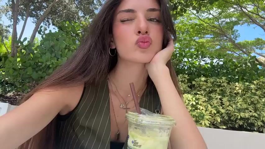 A young woman with long dark hair and light skin is sitting at an outdoor table, leaning her head on her hand. She is wearing a dark green vest over a black top and several necklaces. In front of her is a plastic cup with a straw, containing a green beverage. The background is a blur of green foliage and a bright blue sky.