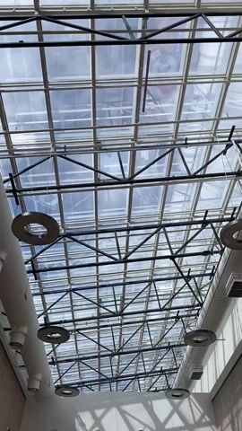 The video shows an upward-facing view of a large glass ceiling with a black metal grid structure supporting it. Sunlight filters through the glass, creating reflections and shadows. Several circular metallic objects and white cylindrical pipes are suspended from the ceiling structure. The scene appears to be inside a building, possibly a mall or an atrium, given the scale and the reflective nature of the objects. No people are visible in the frame.
