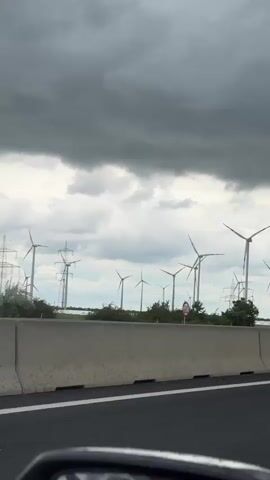 The video, likely taken from a moving vehicle, shows a view of wind turbines against a cloudy, overcast sky. The turbines are located in a field beyond a highway barrier. A speed limit sign reading "80" is visible, along with power lines and street lamps.