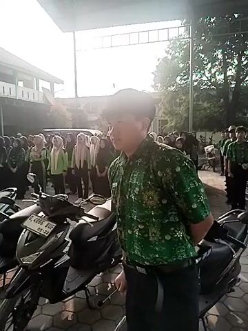 A young person in a green patterned shirt and dark pants stands facing to the left, with their back to the camera, in what appears to be an outdoor setting with a covered walkway overhead. They are holding a white object in their right hand. In front of them, to the left, are several parked motorcycles. Behind them, a large group of people, mostly young women wearing hijabs, are standing in formation, facing the same direction as the person in the foreground. They are all wearing green uniforms. The overall impression is of a school gathering or assembly. The lighting suggests it is daytime, with sunlight filtering in from the right.