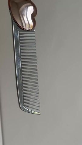 The video shows a metallic comb with a heart-shaped handle. The comb has closely spaced teeth and a reflective surface. The background is a light grey or white wall. The comb is suspended in the air, presumably being held, although the person holding it is not visible. The camera angle and lighting suggest that the video was taken indoors.
