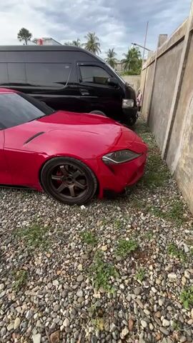 The video shows a red Toyota Supra parked on gravel next to a black van. The Supra has dark tinted windows, dark bronze-colored rims, and aftermarket side skirts. It is parked close to a cement wall. The sky is overcast. There are no people visible in the video.