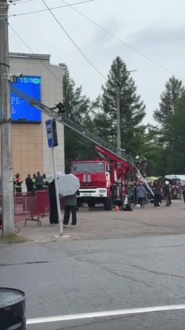 A red fire truck with an extended ladder is positioned in front of a tan building. A firefighter is visible on the ladder, appearing to be working on something attached to a pole with a digital display that shows "15 C". Several other individuals, some in what appear to be safety vests, are standing near the fire truck and a red barrier. Two people are holding umbrellas, suggesting it may be raining. The scene appears to be outdoors, with trees visible in the background and a street in the foreground.