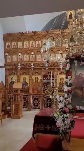 The video pans across the interior of what appears to be an Eastern Orthodox church. In the foreground, a red carpet leads towards a central altar area. To the left, a wooden pulpit stands with a book on it. The main focal point is a large iconostasis, a wall of icons, made of intricately carved wood, adorned with numerous religious icons of saints and religious figures. A large, ornate chandelier hangs from the ceiling. In the center and to the right of the iconostasis, two large framed icons are prominently displayed, decorated with garlands of flowers and greenery. The icon on the right depicts a figure in religious vestments. The overall atmosphere is one of solemnity and reverence, typical of a religious setting.