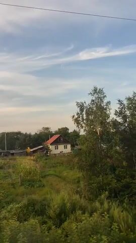 The video shows a view of a rural landscape, likely from a moving vehicle, possibly a train. In the foreground, there are green bushes and trees. In the midground, there are several houses with pitched roofs, gardens, and some farm structures. The houses appear to be in a village or small settlement. The background consists of more trees and a blue sky with scattered white clouds. Power lines are visible in the upper portion of the frame. The overall impression is of a calm, daytime scene in the countryside. The video is short and appears to be a static shot or a very slow pan.