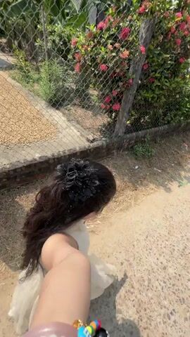 The video, filmed from a first-person perspective, shows a person walking outdoors, holding a selfie stick with a phone. The person has long, dark, wavy hair styled with a black, ornate hair clip resembling flowers or butterflies. They are wearing a light-colored (possibly cream or beige) sleeveless top or dress. On their right wrist, a colorful beaded bracelet is visible, featuring a black bead with a white number "8."

The person is walking on a rustic path that appears to be a mix of dirt and gravel. To their left, a chain-link fence runs parallel to the path, separating it from a vibrant green bush adorned with clusters of bright pink-red flowers, likely Ixora (Jungle Geranium). A low concrete wall is visible at the base of the fence.

In the background, behind the fence and vegetation, several simple residential houses can be seen. Some feature corrugated metal roofs, while others have traditional reddish-brown tiled roofs, characteristic of rural architecture in Vietnam. The scene is brightly lit by direct sunlight, indicating a clear day. Upbeat pop or dance music plays throughout the video. The setting suggests a rural or semi-rural area within Tay Son Commune, Vietnam.