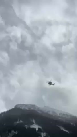 A helicopter flies against a backdrop of a cloudy sky and a mountain with snowy patches. The helicopter is light in color, possibly white or gray, and the mountain is dark, likely due to forestation and shadow. The sky is overcast with layers of gray and white clouds.