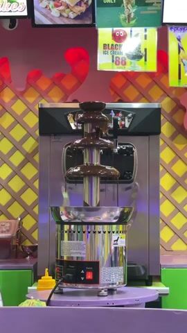 A chocolate fountain is shown in operation in an indoor setting. The fountain is silver with three tiers, and molten chocolate cascades down the sides. The fountain is situated on a purple surface, with yellow and pink decorations visible in the background. A sign advertising "BLACK ICE CREAM 88" is also visible. The lighting is bright and appears to be artificial.