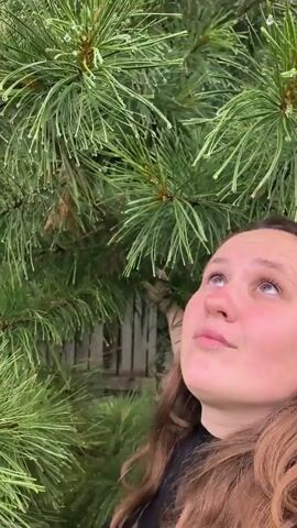 A close-up video shows a young woman with long brown hair standing in front of a pine tree. She has her eyes closed and her head tilted back, seemingly enjoying the rain. As the video progresses, her facial expression changes to one of surprise and laughter. She appears to be reacting to something, possibly a splash of water or a tickle. The pine needles are wet, indicating that it is raining. The background is blurred, with some suggestion of a fence behind the woman.