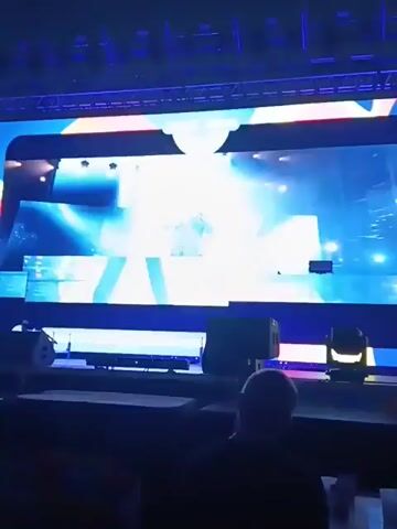 From a low angle, a brightly illuminated large screen displays video footage of what appears to be a concert or performance. At times, the screen displays a person performing, alternating with aerial shots of a large audience. Below the screen, stage lighting equipment and speakers can be seen. Two people are seated in front of the camera. One person appears to be bald, and the other is wearing a camouflage patterned shirt. The word "Eurovision" is displayed at the top of the screen.
