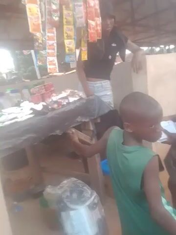 This is a poorly lit, vertically oriented video of a stall with various items for sale. In the foreground, a young boy in a green tank top is reaching for something above him. Behind him, another child in a black shirt and striped shorts is leaning over a table. The stall appears to be outdoors, with a corrugated metal roof visible. Stacks of packaged goods, possibly food items, are displayed on shelves. There are also plastic cups bundled together. The overall impression is of a small, informal market or shop.