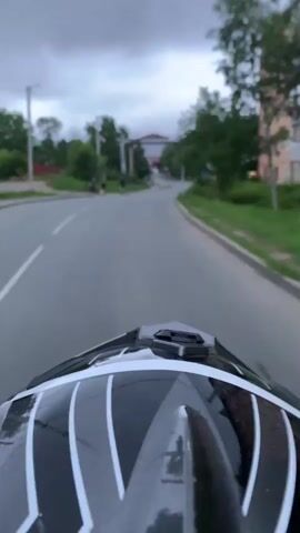 This media file is a first-person perspective video, likely from a helmet camera mounted on a motorcycle or similar vehicle. The subject is a person riding a vehicle on a paved road. The scene is an urban or suburban environment with buildings and trees visible.

**Subjects:** The primary subject is the rider's helmet, which is black with white stripes and a black visor. In the background, there are several people walking on the sidewalk, a teal car parked on the side of the road, and a red car further down the road. Buildings, including a multi-story brick building, are visible. Trees and green foliage line the road. A road sign with a yellow diamond shape is also visible.

**Activity:** The person is riding a vehicle along a road. The road appears to be a public street, with lanes marked by white lines.

**Notable Details:**
*   **Time of Day:** The lighting suggests it is either early morning or late afternoon, with a cloudy sky.
*   **Weather:** The sky is overcast, indicating a cloudy or possibly impending rain.
*   **Location Cues:** The presence of apartment buildings, trees, and road infrastructure are typical of a developed area. The specific mention of Artyom, Russia, provides geographical context.
*   **Emotions:** The video does not explicitly convey emotions, but the first-person perspective creates a sense of immersion and movement.

**Visible Text:** No significant or legible text is visible in the video.