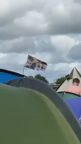 Here's a description of the video:

The video shows a festival campsite with several tents visible in the foreground. The tents are of various colors including green, grey, and blue. In the background, a flag is waving. The flag features a collage of images of four people. The sky is overcast with grey and white clouds. A man is standing shirtless near one of the tents.