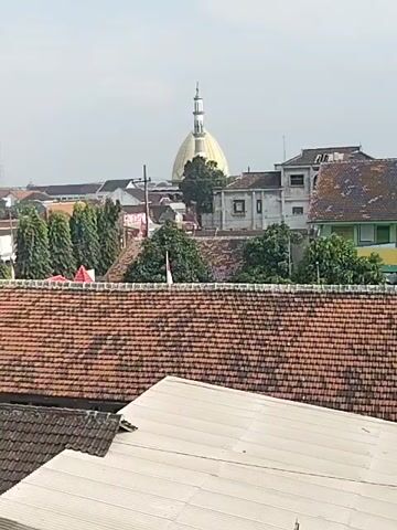 This is a static, aerial view of a cityscape. In the foreground, there are several rooftops, the closest of which are made of reddish-brown tiles, and another of a lighter, possibly metal material. Behind these rooftops, a line of tall, thin evergreen trees is visible. In the middle ground, various buildings are present, including structures with white and grey walls. A prominent, large, golden dome with a minaret topping it is visible in the background, suggesting the presence of a mosque or similar religious building. An Indonesian flag is seen flying on a pole in front of some trees. The sky is overcast.