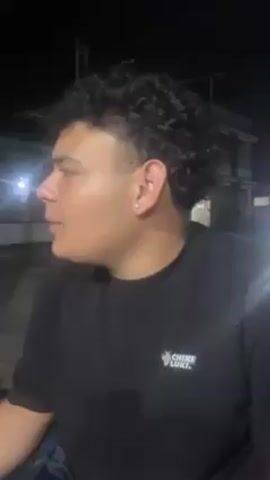 Here's a description of the video:

The video shows a young man speaking, with his face partially visible. He has curly dark hair and a light skin tone. He is wearing a black t-shirt with a small logo on it. The background is dark, suggesting it is night, with some buildings and lights visible in the distance. The camera then quickly pans to show a dark, indistinct figure moving in the background before turning back to focus on the young man again. The lighting is poor and makes it difficult to make out fine details.