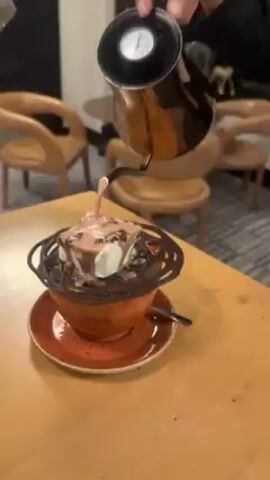 Here is a description of the video:

A hand pours chocolate sauce from a silver kettle over a cup of hot chocolate. The hot chocolate has a marshmallow and some chocolate shavings on top, and a chocolate rim that looks like a nest. The cup sits on a matching saucer, and a spoon rests nearby. The background shows a wooden table, chairs, and part of the room's interior. The kettle has a thermometer on top, indicating the liquid's temperature.