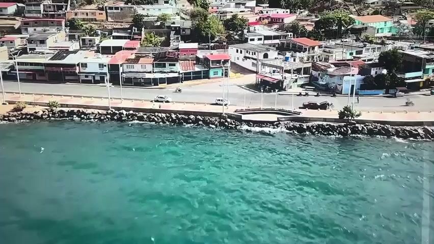 This media file is a video showcasing a coastal town. The first scene, timestamped 00:00, provides an aerial view of a seaside town with buildings lining a waterfront promenade. A road runs along the promenade, with a few cars and motorcycles visible. The buildings are a mix of residential and commercial structures, painted in various colors. The sea is a vibrant turquoise, with gentle waves lapping against the shore. The scene appears to be set during the daytime, with clear skies and bright sunlight.

The subsequent scenes, from 00:01 to 00:04, transition to a wide shot of a vast expanse of blue ocean under a clear sky. The water is a deep blue, transitioning to lighter, greenish hues closer to the shore, suggesting shallow depths. In the distance, a hazy mountain range forms the horizon. The sky is a brilliant, cloudless blue, indicating fair weather. The overall mood is serene and expansive, highlighting the natural beauty of the coastal environment.

While the prompt mentions Valladolid, Spain as a location context, the visuals do not contain any explicit cues or landmarks that definitively identify it as such. The architecture and the general setting are more indicative of a tropical or Mediterranean coastal region. There is no visible text within the video. The media appears to capture peaceful daytime scenes of a coastal town and the adjacent sea.