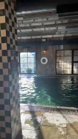 The video shows an indoor pool with a tiled column in the foreground. The column has a black and white checkerboard pattern. The pool itself is a light green color and the water is rippling. Beyond the pool, there are two large windows allowing sunlight to filter into the room. Between the windows, a white life preserver hangs on the wall. The wall is made of wooden planks. The ceiling is composed of a series of wooden beams or planks.