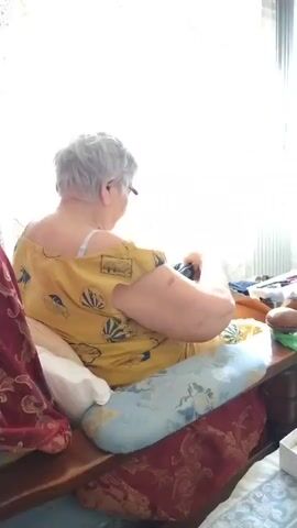 An elderly woman with gray hair and glasses is sitting on a wooden bench, facing away from the camera. She is wearing a yellow shirt with a pattern of hot air balloons and light blue patterned pants. She is holding a dark blue object in her hands and appears to be folding or manipulating it. To her right, on the bench, is a brown oval object, a green plastic bag, and other items. Behind her, a window is covered with a light-colored, decorative curtain, and a radiator is visible to the right. The scene is indoors, with natural light coming from the window.