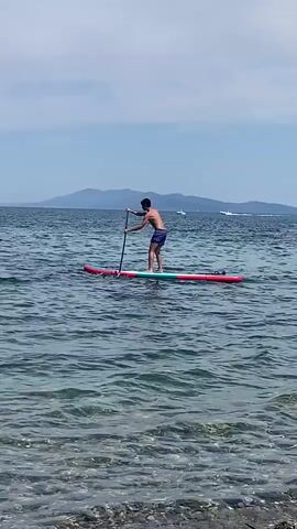 A man is paddleboarding on a calm sea under a cloudy sky. He is wearing blue shorts and is holding a paddle. A woman is swimming in the foreground, facing away from the camera. In the distance, there are islands and boats. The water is clear and the seabed is visible in the foreground.