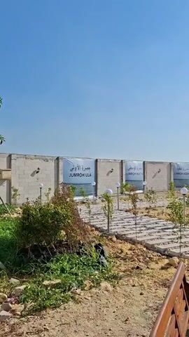 The video shows an outdoor area under a clear blue sky. In the foreground, there is a sandy area with some foliage and a wooden bench. A paved walkway with square stepping stones leads into the scene. Along the walkway, there are young trees and lamps. In the background, a concrete wall runs horizontally, with signs posted on it. The wall is topped with what appears to be a fence. Beyond the wall, there are some buildings and distant hills. A large, white tensile structure provides shade over a paved area. A pillar decorated with ornate carvings supports the structure. Several motorcycles are parked in front of a building that resembles the Kaaba in Mecca, with black walls and gold accents. The overall impression is of an outdoor recreational or religious site.