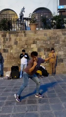 A man in a brown shirt and jeans dances and raps into a microphone in a public area. He is positioned in front of a stone brick wall, where two other young men stand, watching him. One of the men is recording with his phone. A speaker and some bags are placed near them. Behind the wall is a platform with a statue of a donkey. Several people gather to watch the performance.