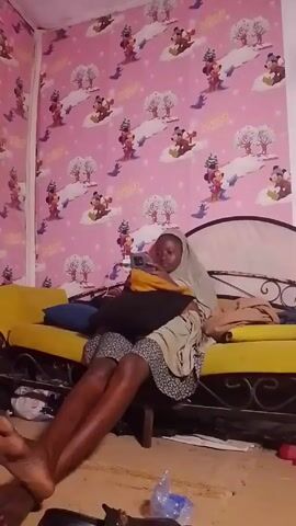 The video shows a person sitting on a yellow bed frame with a white headboard, in a room with pink Disney-themed wallpaper. The person appears to be using a mobile phone. They are wearing a beige headscarf, a patterned dress, and appear to be leaning against a black pillow. Under the bed frame, there are items, including a pair of black shoes, a plastic bag, and what seems to be a white sock.