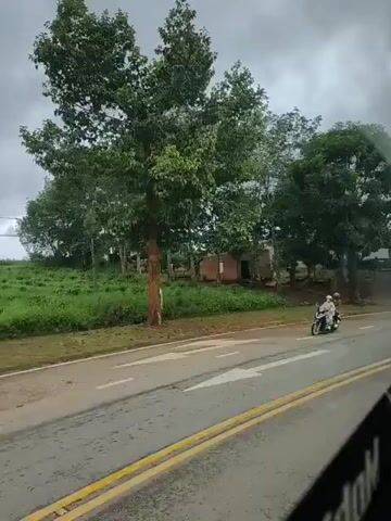 The video shows a road scene from the perspective of a vehicle moving along it. A gray, cloudy sky is visible. On the left side of the road, there are trees and vegetation. In the foreground, a motorcycle with two occupants is traveling on the wet asphalt road, which has yellow double lines marking the lanes. The road appears to be in a rural or semi-rural setting, with a small building visible in the distance on the left. The environment suggests it might be raining or has recently rained.