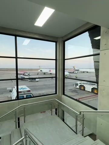 The video is a static shot taken from inside an airport terminal, looking out onto the runway. Large windows offer a clear view of several parked airplanes, including one with a red and white livery and the name "Eurowings" on its side. Other planes can be seen, one with a green tail. Airport service vehicles, including an ambulance, are visible on the tarmac. A few people in high-visibility vests are working around the aircraft. The light suggests it's either early morning or late afternoon, as the sky has a soft, faded color. In the foreground, there's a staircase with stainless steel railings and concrete steps. The interior is modern with a minimalist design, featuring concrete walls and bright overhead lighting.