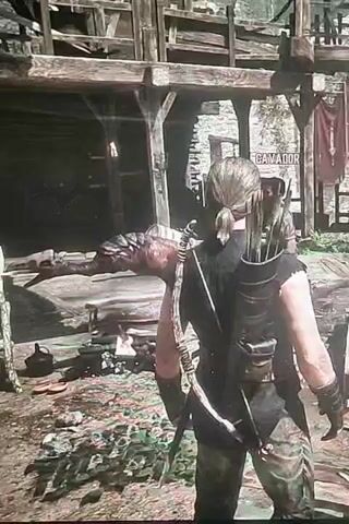 A video shows a person with a bow and quiver on their back, standing in front of a wooden structure. The person is facing away from the camera. The structure appears to be an outdoor market or village. There are barrels and other objects scattered around the ground. A sign that reads "CAMADOR" is visible on the structure. The person's attire suggests a medieval or fantasy setting. The video is short and shows no action or interaction.