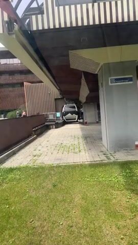 A cable car is being maneuvered into a station. The camera appears to be inside another cable car that is stationary. The cable car in view is white with black accents and large windows. It is positioned on a track within the station. The station appears to be made of modern materials, with metal and concrete elements. There is a paved area in front of the station with some sparse vegetation growing through the cracks. The lighting suggests it is daytime.