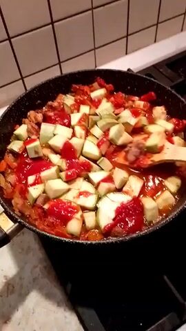 A close-up shot shows a black frying pan on a stovetop containing a mixture of ingredients being stirred with a wooden spoon. The mixture includes diced zucchini, a red sauce resembling tomato sauce, and pieces of meat. The stovetop is black and located near a wall covered with white, square tiles with dark grout. A person's hand is visible holding the wooden spoon.