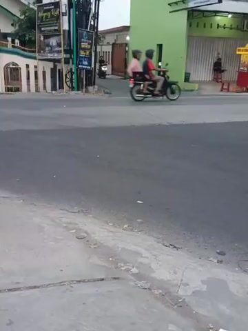 The video shows a street scene. On the left, there is a light-colored building with a green trim and a fence. There are several motorcycles parked in front of the building. A street lamp with signs is located in the middle of the frame. To the right, there is a bright green building. Two people are riding on a motorcycle on the road, traveling from right to left. Another person is riding a motorcycle on the road, traveling from left to right. The road surface is asphalt. The area appears to be an urban or suburban environment.