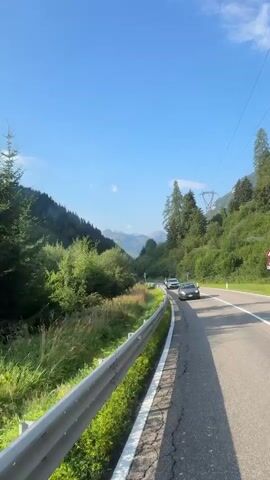 This is a vertical, first-person perspective video taken from a vehicle traveling along a paved road. The road is bordered on the left by a metal guardrail and a grassy embankment leading down to a stream or river. Beyond the embankment are dense green trees and vegetation. In the distance, a valley opens up with more trees and mountains. A bright blue sky with a few wispy clouds is visible. Several vehicles are on the road ahead: a white van, a dark sedan, and a white bus. The sun appears to be shining, casting shadows on the road. Power lines are visible in the upper right portion of the frame.