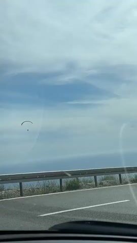 A paraglider is seen in the sky, with a vast ocean and rolling hills in the background. A highway runs along the edge of a cliff, with a metal guardrail separating the road from the drop-off. The sky is partly cloudy, with patches of blue sky visible. The video appears to be shot from a moving vehicle, as indicated by the slight blur and the perspective of the road. The paraglider is small in the frame, suggesting it is some distance away, and it is captured against the backdrop of the ocean and sky. The overall scene suggests a scenic coastal drive.